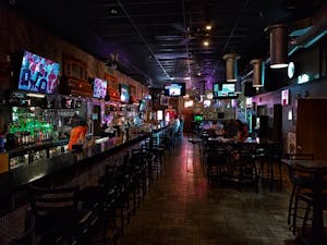 A dimly lit modern bar interior featuring a long counter and multiple screens, creating a cozy urban atmosphere.